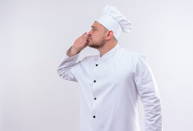 Young handsome cook in chef uniform doing tasty gesture looking at side on isolated white space