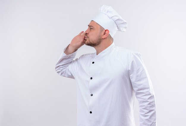 Молодой красивый повар в униформе шеф-повара делает вкусный жест, глядя в сторону на изолированном белом пространстве