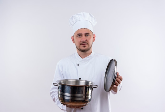 Young handsome cook in chef uniform holding boiler and its cover looking  on isolated white space 