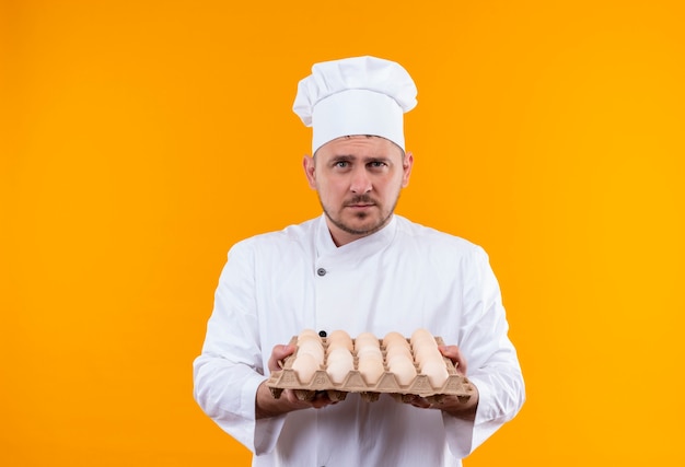 Young handsome cook in chef uniform holding carton of eggs looking  isolated on orange space