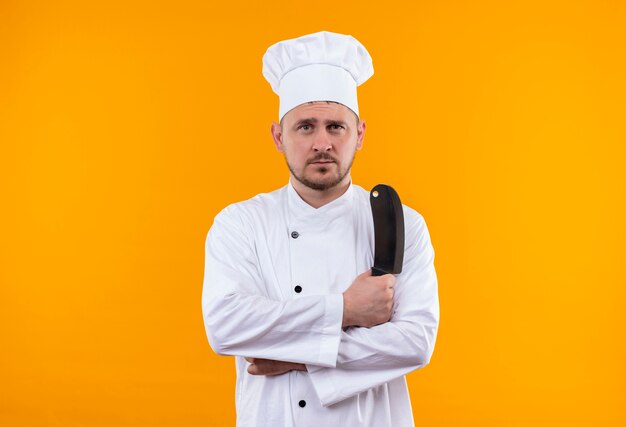 Young handsome cook in chef uniform holding cleaver looking  isolated on orange space