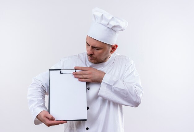 クリップボードを保持し、孤立した白いスペースでそれを見てシェフの制服を着た若いハンサムな料理人