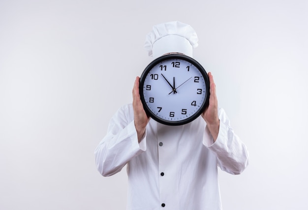 Free photo young handsome cook in chef uniform holding clock and hiding behind it isolated on white space