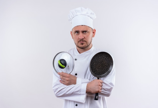 Young handsome cook in chef uniform holding frying pan and pan lid looking  isolated on white space 