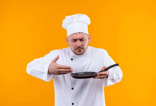 Young handsome cook in chef uniform holding frying pan and sniffing isolated on orange space