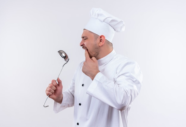 Молодой красивый повар в униформе шеф-повара держит ковш, глядя на него и кладет палец в рот на изолированном белом пространстве