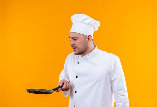 Young handsome cook in chef uniform holding and looking at frying pan isolated on orange space