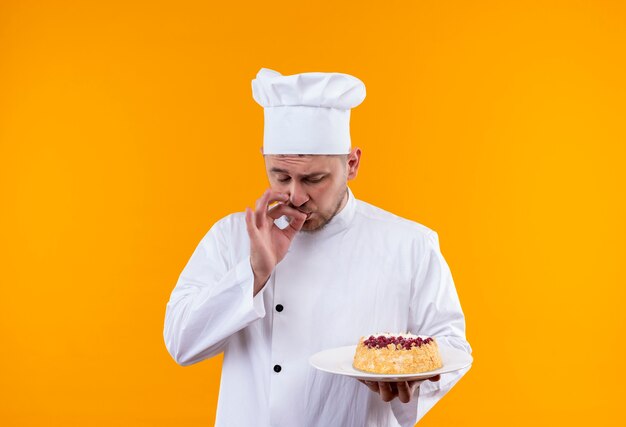 Молодой красивый повар в униформе шеф-повара держит тарелку торта и делает вкусный жест, изолированный на оранжевом пространстве
