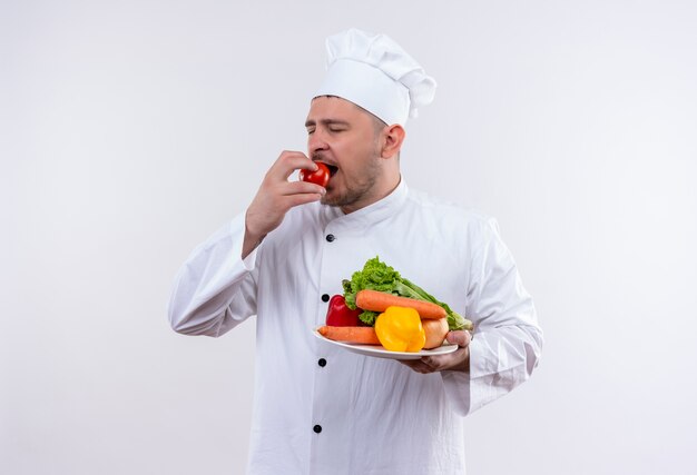 Young handsome cook in chef uniform holding plate with vegetables and biting tomato with closed eyes on isolated white space