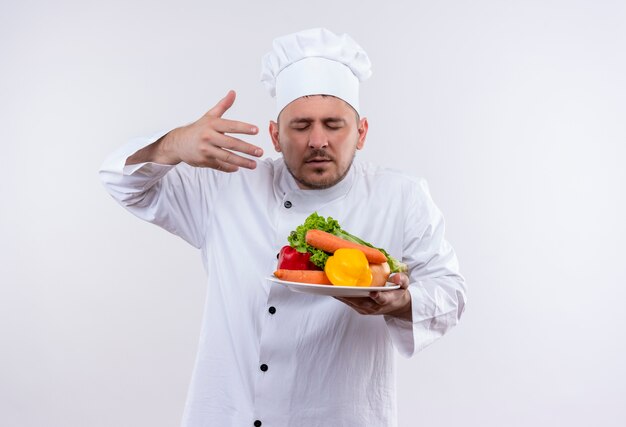 Young handsome cook in chef uniform holding plate with vegetables sniffing them with raised hand and closed eyes on isolated white space