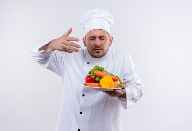 Молодой красивый повар в униформе шеф-повара держит тарелку с овощами, обнюхивая их поднятой рукой и закрытыми глазами на изолированном белом пространстве