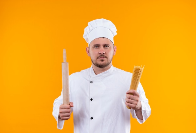 Молодой красивый повар в униформе шеф-повара держит скалку и макароны для спагетти, глядя изолированно на оранжевом пространстве