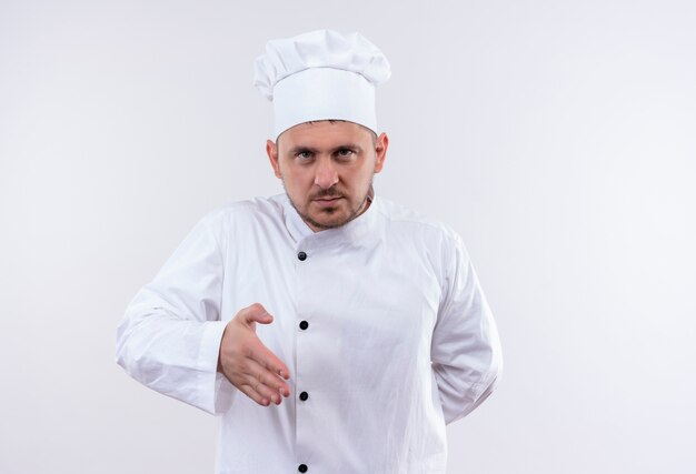 Young handsome cook in chef uniform stretching out hand gesturing hi with another hand behind his back looking  on isolated white space