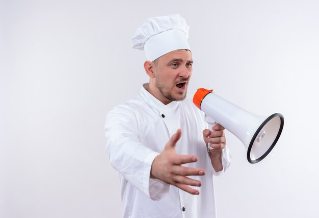 Young handsome cook in chef uniform talking by speaker and stretching out hand isolated on white space