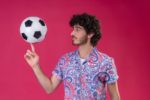 Young handsome curly man holding soccer ball on finger and looking at it on isolated pink wall