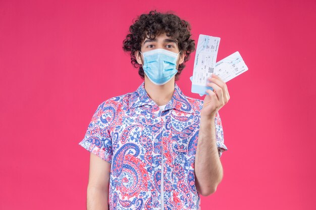 Молодой красивый кудрявый путешественник в медицинской маске держит билеты на самолет на изолированной розовой стене с копией пространства
