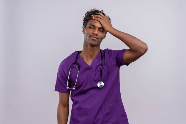 A young handsome dark-skinned doctor with curly hair wearing violet uniform with stethoscope feeling bad having high temperature physical pressure keeping eyes closed touching forehead with pa