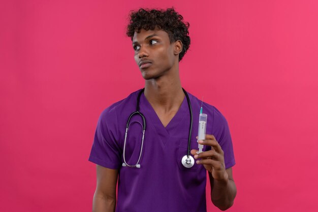 A young handsome dark-skinned doctor with curly hair wearing violet uniform with stethoscope holding injection syringe 