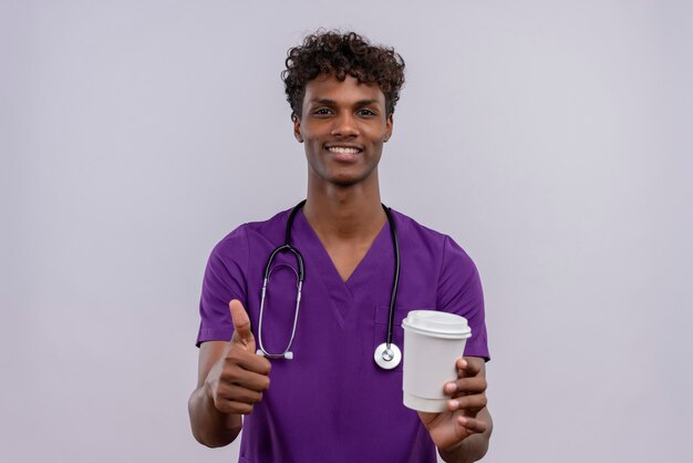 Молодой красивый темнокожий врач с кудрявыми волосами в фиолетовой форме со стетоскопом держит бумажный стаканчик с кофе с большими пальцами руки вверх