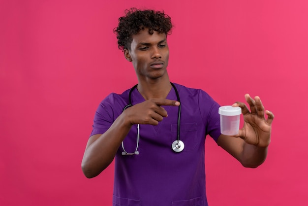 A young handsome dark-skinned doctor with curly hair wearing violet uniform with stethoscope pointing with index finger at medical plastic specimen jar 