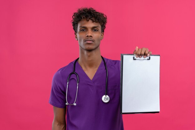 A young handsome dark-skinned doctor with curly hair wearing violet uniform with stethoscope showing clipboard with blank sheet of paper 