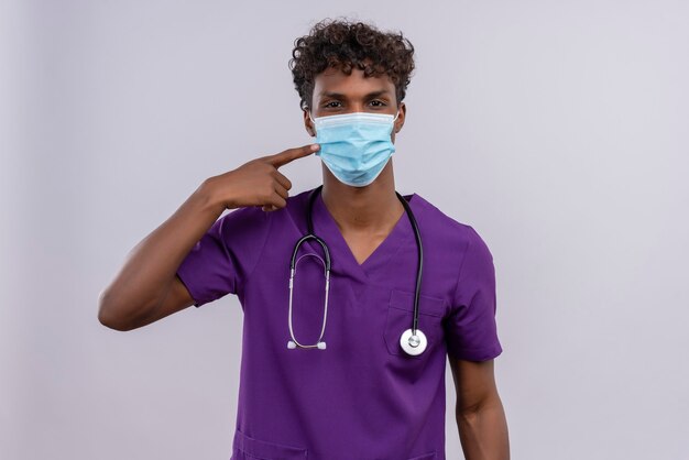 A young handsome dark-skinned doctor with curly hair wearing violet uniform with stethoscope showing face mask 