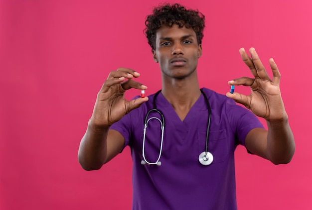 Молодой красивый темнокожий врач с кудрявыми волосами в фиолетовой форме со стетоскопом показывает таблетки
