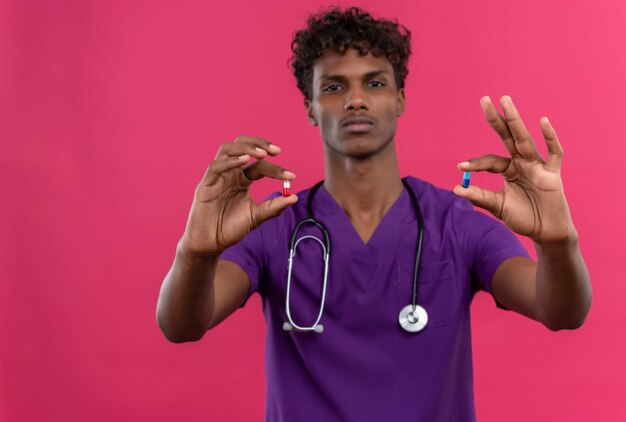 A young handsome dark-skinned doctor with curly hair wearing violet uniform with stethoscope showing pills 