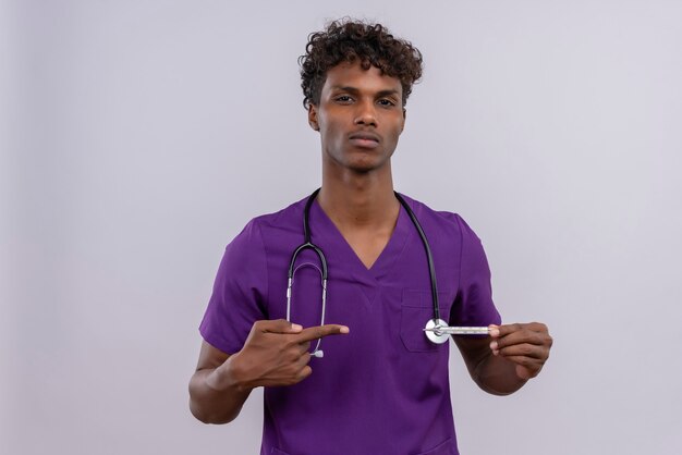 Молодой красивый темнокожий врач с кудрявыми волосами в фиолетовой форме со стетоскопом показывает большие пальцы вниз, указывая указательным пальцем на термометр