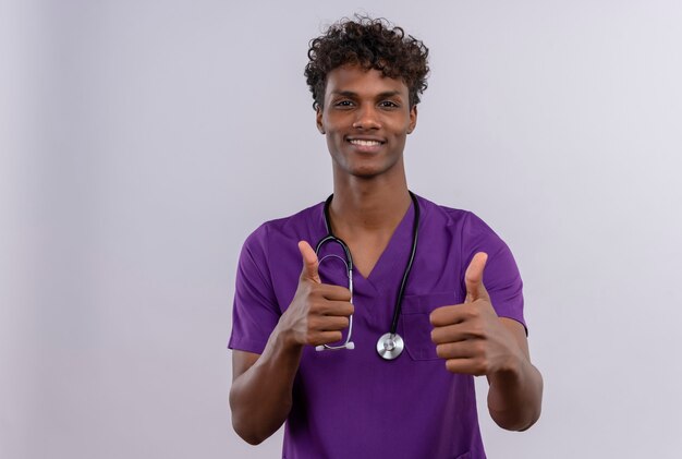 A young handsome dark-skinned doctor with curly hair wearing violet uniform with stethoscope showing thumbs up with happy face 