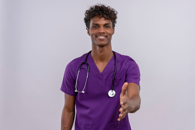 Молодой красивый темнокожий врач с кудрявыми волосами в фиолетовой форме со стетоскопом протягивает руку для рукопожатия, чтобы поприветствовать кого-то или поздороваться