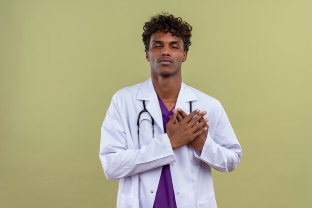 A young handsome dark-skinned doctor with curly hair wearing white coat with stethoscope feeling ache while touching heart on a green space