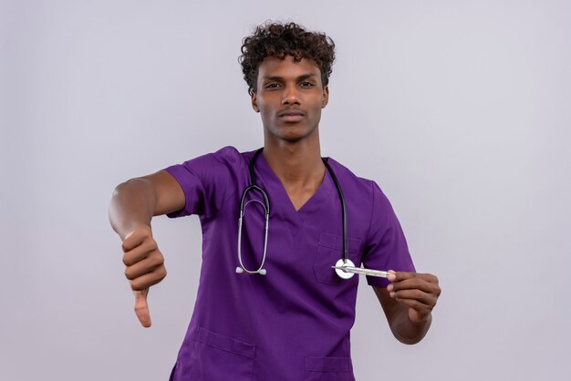 A young handsome dark-skinned male doctor with curly hair wearing violet uniform with stethoscope showing thumbs down while holding thermometer 