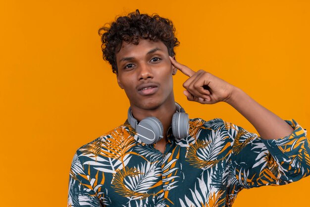 Free photo a young handsome dark-skinned man with curly hair in leaves printed shirt in headphones pointing with index finger on head