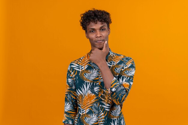 A young handsome dark-skinned man with curly hair in leaves printed shirt keeping hand on chin 