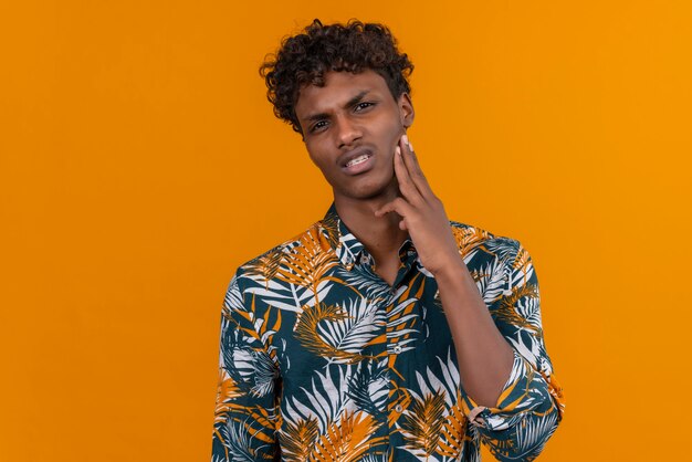 A young handsome dark-skinned man with curly hair in leaves printed shirt with angry face with hands on cheek 