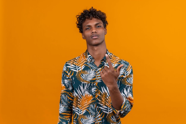 A young handsome dark-skinned man with curly hair in leaves printed shirt with confident expression raising hand 