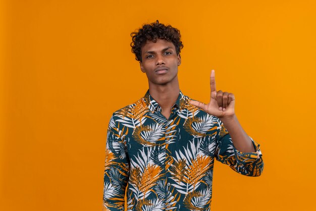 A young handsome dark-skinned man with curly hair in leaves printed shirt with serious face raising hand 