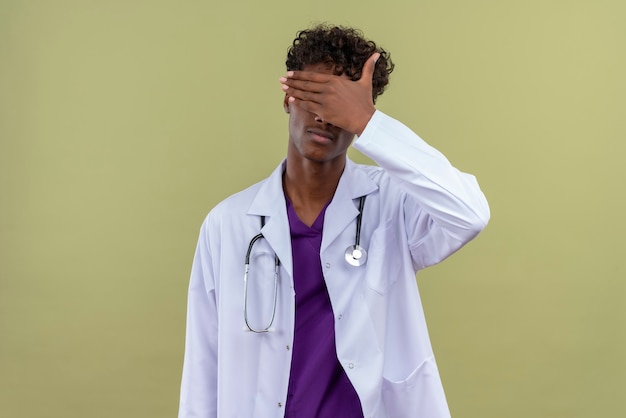 A young handsome dark-skinned man with curly hair wearing white coat with stethoscope closing his eyes with palm on a green space