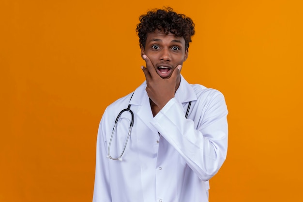 A young handsome dark-skinned man with curly hair wearing white coat with stethoscope looking surprisingly at camera 