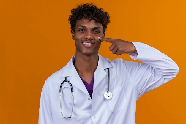 Free photo a young handsome dark-skinned man with curly hair wearing white coat with stethoscope pointing with index finger on cheek