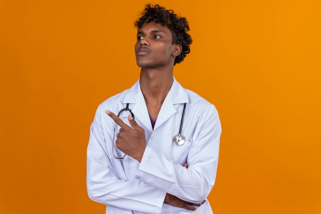 A young handsome dark-skinned man with curly hair wearing white coat with stethoscope pointing with index finger 