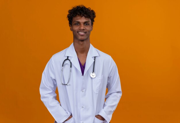 A young handsome dark-skinned man with curly hair wearing white coat with stethoscope putting hands on pockets 