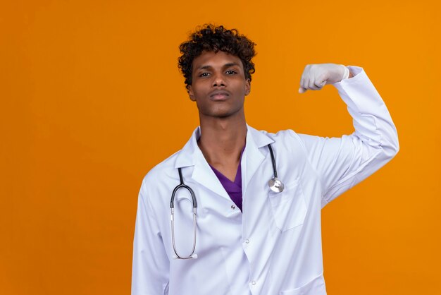 A young handsome dark-skinned man with curly hair wearing white coat with stethoscope raising hand with clenched fist 