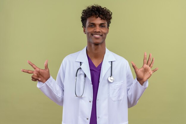 Free photo a young handsome dark-skinned man with curly hair wearing white coat with stethoscope showing number eight on a green space