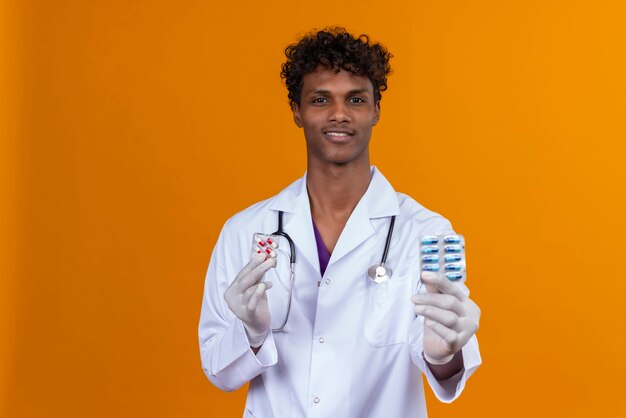 A young handsome dark-skinned man with curly hair wearing white coat with stethoscope showing pills 