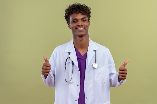 A young handsome dark-skinned man with curly hair wearing white coat with stethoscope smiling while showing thumbs up on a green space