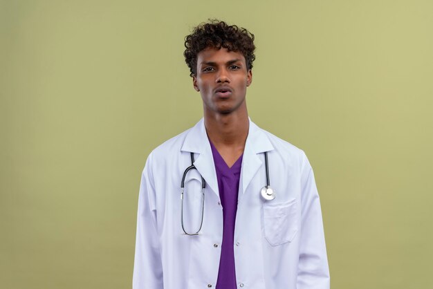 A young handsome dark-skinned man with curly hair wearing white coat with stethoscope  surprisingly on a green space