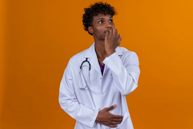 A young handsome dark-skinned man with curly hair wearing white coat with stethoscope thinking with hands on mouth 