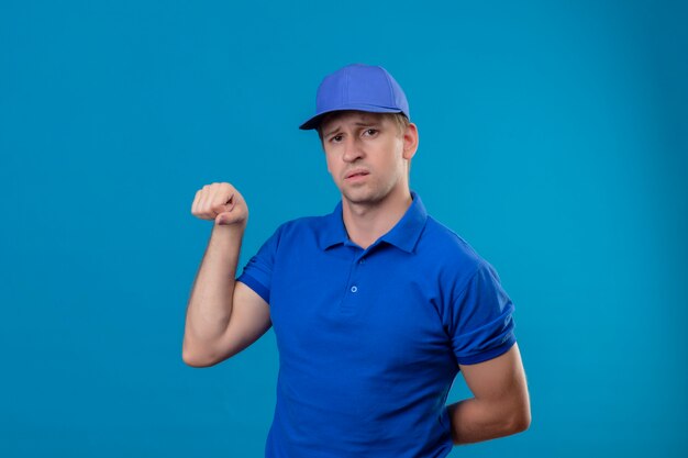 Young handsome delivery man in blue uniform and cap clenching fist showing biceps looking confident standing over blue wall